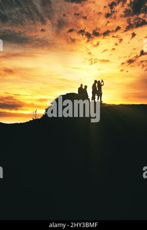 Silhouette von vier Personen, die auf dem Gipfel des Berges auf dem goldenen Hintergrund des Sonnenaufgangs fotografieren Stockfoto