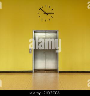 Silberne Aufzugstür mit der Uhr oben im Gebäude mit gelben Wänden Stockfoto