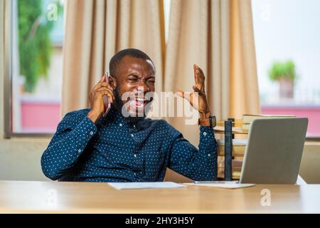 Der junge Schwarze aus Ghana, der auf seinem Schreibtisch arbeitet und wütend am Telefon schreit Stockfoto