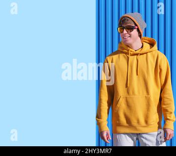 Fröhlicher junger Mann in einem gelben Sweatshirt und einem Wollhut, der auf den blauen Himmel mit Wolken zeigt Stockfoto