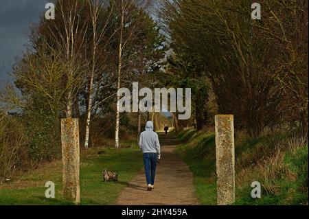 Teenager-Junge in grauem Kapuzenoberteil und Jeans, der unter stürmischem Himmel den Cairn Terrier-Hund entlang des Stratford Greenway führt Stockfoto