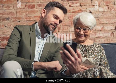 Erwachsener kaukasischer Sohn zeigt seiner älteren grauhaarigen Mutter etwas auf seinem Smartphone, während er auf einem Sofa sitzt. Hochwertige Fotos Stockfoto