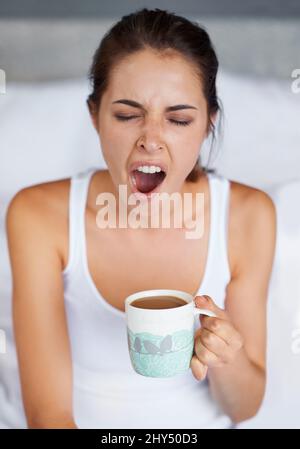 Ein großes GÄHNEN. Eine junge Frau, die ein großes Gähnen gibt, während sie mit einer Tasse Kaffee im Bett sitzt. Stockfoto