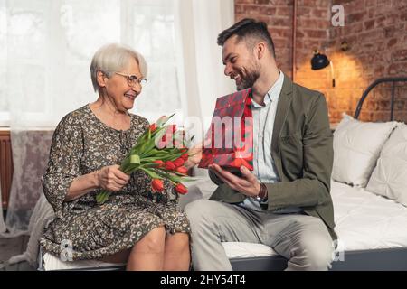 kaukasischer Sohn gibt seiner Mutter rote Tulpen und rote Geschenktüte zum Muttertag. Hochwertige Fotos Stockfoto