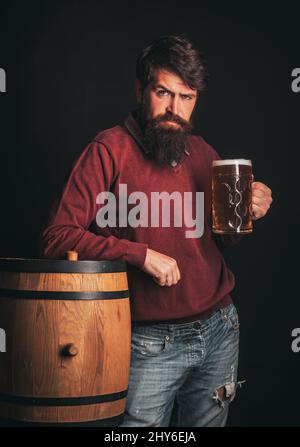 Älterer Mann trinkt Bier mit Überraschungsgesicht. Brauer. Stilvoller hübscher Mann, der Bier auf schwarzem Hintergrund trinkt. Mann mit Bart mit Holzfass und Stockfoto