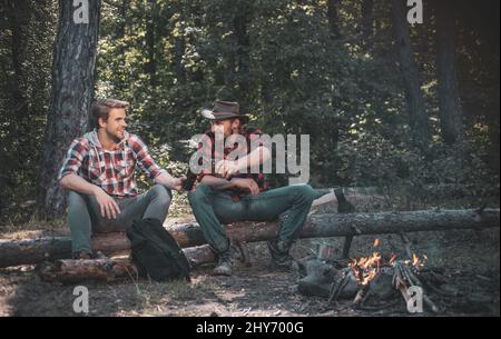 Zwei gutaussehende Männer Freunde auf Camping in der Nähe Lagerfeuer. Touristen entspannen sich. Zwei Freunde Holzfäller sitzen im Wald mit Bier. Freunde genießen es Stockfoto