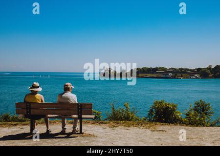 Seniorenpaar sitzt auf einer Holzbank vor dem Meer auf einem blauen Himmel Hintergrund Stockfoto