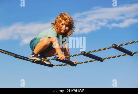 Netter Junge klettert die Leiter auf dem Spielplatz hoch. Kind klettert die Leiter gegen den blauen Himmel hinauf. Schöner lächelnder netter Junge auf einem Spielplatz. Kinder Stockfoto