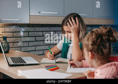 Mutter benutzte einen Laptop, um ihrer Tochter bei den Hausaufgaben zu helfen, die sie am Tisch in der Küche saß. Heimschooling. Stockfoto