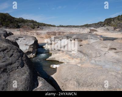 Der Pedernales River im Pedernales Falls State Park in der Nähe von Johnson City, Texas. Ein kleiner Wasserfall aus Kalkstein ist im Vordergrund und zwei Wanderungen Stockfoto