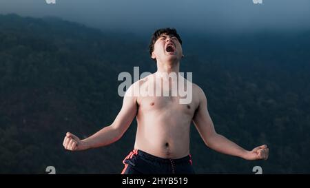 Schöner Mann ohne Hemd, der auf dem Gipfel des Berges mit weißem Nebel steht. Erfolgreicher Mann Wanderer öffnen die Arme und genießen den wunderschönen Sonnenaufgang in der Morgendämmerung Stockfoto