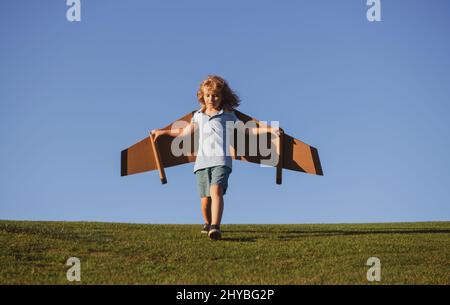 Kind, das im Flugzeug fliegt, machte Handwerk aus Pappflügeln. Traum, Phantasie, Kindheit. Reise- und Sommerurlaubskonzept. Kleiner Junge Pilot gegen einen blauen Stockfoto