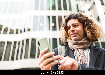 Von Männern unter mittlerem Alter mit lockigen Haaren, die auf dem Handy in sozialen Medien surfen, während sie gegen ein modernes Gebäude auf der Stadtstraße stehen Stockfoto