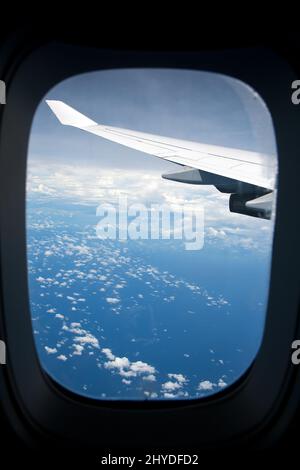 Fliegen über den Wolken. Blauer Himmel, Wolken und Flugzeugflügel über dem Ozean an einem sonnigen Tag durch ein Fenster gesehen. Stockfoto