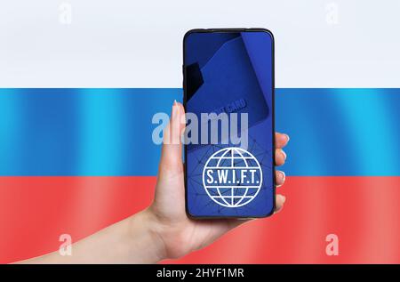 Die Hand einer Frau hält ein Mobiltelefon. Auf dem Smartphone-Bildschirm Zeichen von S.W.I.F.T. Speicherplatz kopieren. Die russische Flagge ist im Hintergrund. Das Konzept von i Stockfoto