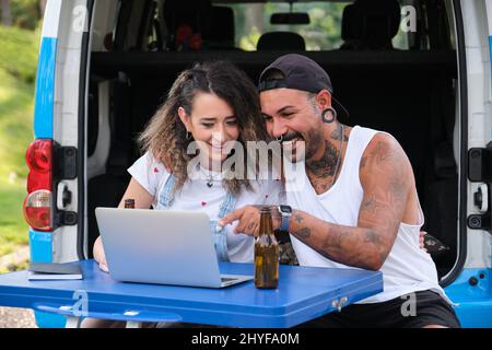 Das junge tätowierte Paar lächelt und sieht etwas im Laptop in der Nähe des Wagens. Stockfoto