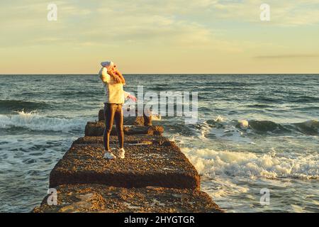 Eine Frau mit blondem Haar in einem Hut und einem Pelzmantel geht an einem Steinbrecher entlang des Meeres Stockfoto