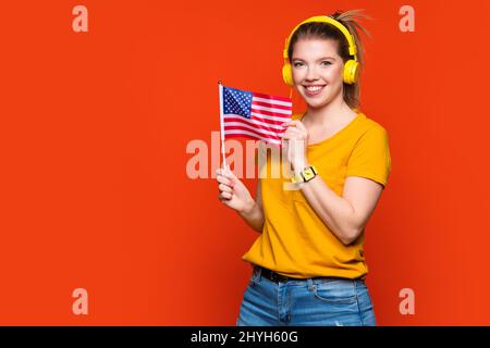 Mädchen in gelbem Grundanzug und Kopfhörern hält eine kleine USA-Flagge. Studieren Sie im Ausland und reisen Sie. Internationales Austauschprogramm für Studenten. Amerikanisches Englisch lernen mit Muttersprachlern. Fremdsprache durch Ohr Stockfoto