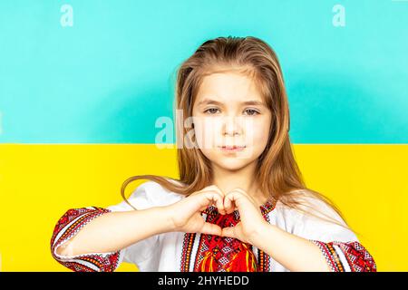 Ein Mädchen in ukrainischer Nationalkleidung, Vyschywanka, zeigt ein Herzzeichen als Zeichen der Liebe zur Ukraine, Nahaufnahme vor dem Hintergrund der Ukrainerin Stockfoto