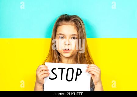 Das Mädchen hält vor dem Hintergrund der ukrainischen Flagge ein STOPPPLAKAT in den Händen. Beendet den Krieg in der Ukraine. Das Konzept des Friedens in der Ukraine Stockfoto