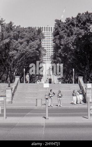 Sydney Australia 1977: Blick vom nördlichen Teil des Hyde Parks über die Park Street auf die Treppe und den zentralen Pfad, der bis zur Liverpool Street in Sydney führt. Im Jahr 2022 ist der zentrale Straßenteiler (Vordergrund) verschwunden und die Bäume füllen nun die Lücke und verstecken so das Gebäude an der Liverpool Street 175, und die Outfits haben sich geändert, aber ansonsten hat sich wenig geändert. Stockfoto