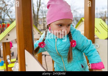 Porträt eines Vorschulkindes auf dem Spielplatz. Ein fünfjähriges Mädchen in einem rosa Hut geht die Treppe hoch und runter und hält sich an das Geländer. Kindheit. Stockfoto