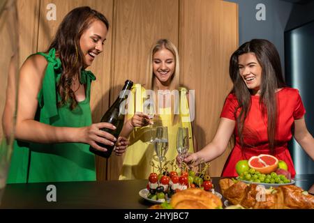 Fröhliche Mädchen, die Champagner während eines freundlichen Treffens zu Hause in die Gläser gießen Stockfoto