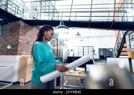 Lächelnde afroamerikanische junge Architektin, die im modernen Büro steht und Blaupause hält Stockfoto