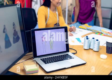 Kleidungsentwürfe auf Laptop und Schreibtisch mit mehrrassischen weiblichen Designerinnen im Hintergrund Stockfoto