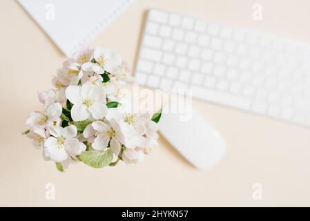 Tastatur, weißes Blatt Papier und Maus und weiße Apfelblüten in einer Vase, die nicht fokussiert auf einem beigen Tisch liegt. Layout- und Designkonzept. Flach liegend. Stockfoto