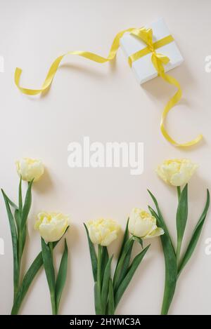 Geschenkschachtel in weißem Papier mit einem gelben Band und Blumen aus gelben Tulpen auf einem hellen Tisch verpackt. Flach liegend. Platz für Text kopieren. Stockfoto