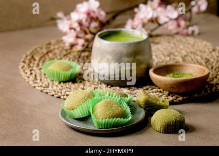 Asiatischer Reis Dessert süßer grüner Matcha-Mochi mit einer Tasse aufgeschäumtem Matcha-Tee und grünem Matcha-Pulver in Keramikplatte auf braunem Tisch. Teekannen und Frühling Stockfoto