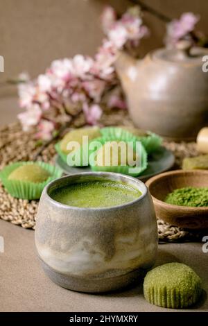 Asiatischer Reis Dessert süßer grüner Matcha-Mochi mit einer Tasse aufgeschäumtem Matcha-Tee und grünem Matcha-Pulver in Keramikplatte auf braunem Tisch. Teekannen und Frühling Stockfoto