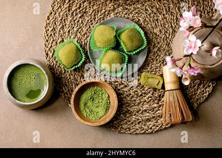 Asiatischer Reis Dessert süßer grüner Matcha-Mochi mit einer Tasse aufgeschäumtem Matcha-Tee und grünem Matcha-Pulver in Keramikplatte auf braunem Tisch. Teekannen und Frühling Stockfoto