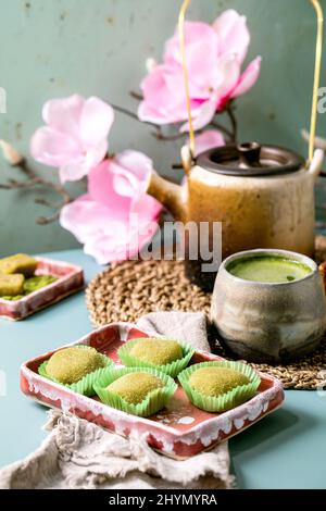 Asiatische traditionelle Reis Dessert süßen grünen Matcha-Mochi in rosa Teller mit einer Tasse aufgeschäumten Matcha-Tee auf blauem Tisch. Alte Teekannen und frühlingshafte rosa Blüten Stockfoto