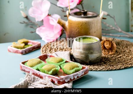 Asiatische traditionelle Reis Dessert süßen grünen Matcha-Mochi in rosa Teller mit einer Tasse aufgeschäumten Matcha-Tee auf blauem Tisch. Alte Teekannen und frühlingshafte rosa Blüten Stockfoto