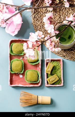 Asiatische traditionelle Reis Dessert süßen grünen Matcha-Mochi in rosa Keramik-Schale mit Tasse aufgeschäumter Matcha-Tee auf blauem Tisch mit Frühling rosa Blüten. Fl Stockfoto