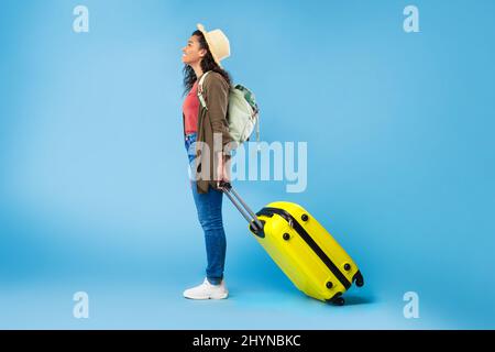 Seitenansicht einer jungen schwarzen Frau mit Rucksack und einem hellen Koffer, die in den Urlaub fährt und ins Ausland reist Stockfoto