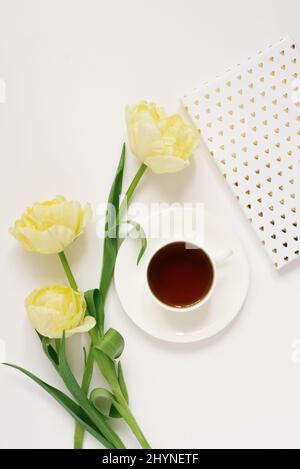 Eine weiße Porzellanschale mit schwarzem Tee auf einer Untertasse, umgeben von frühlingsgelben Tulpen, ein Laptop auf weißem Hintergrund. Das Konzept des Frühlings, Frauen und Stockfoto