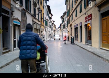 Mann, der jemanden auf einem Rollstuhl in der Mitte einer Straße im historischen Zentrum von Florenz mit einem Auto vor sich trägt Stockfoto