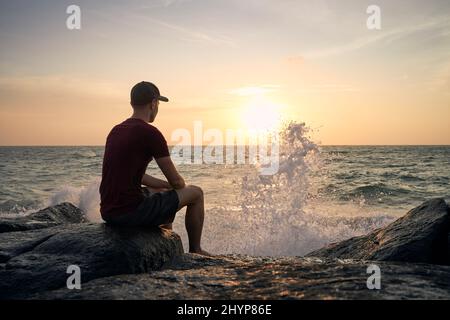 Rückansicht eines Mannes, der auf einem Felsen sitzt und den wunderschönen Sonnenuntergang über dem Meer beobachtet. Stockfoto