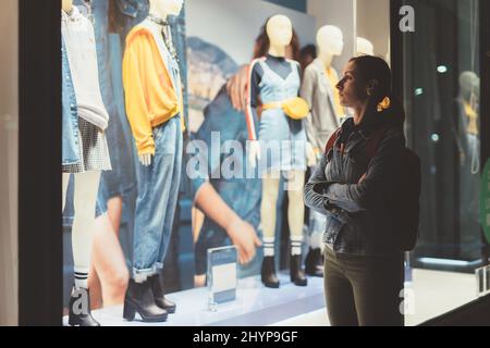 Mädchen steht vor dem Laden und schaut auf die weiblichen Kleider auf den Schaufensterpuppen Stockfoto