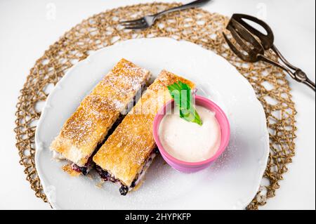2 Stück Kuchen aus Blätterteig mit Heidelbeere und Creme, kleine rosafarbene Schüssel mit Creme, Gabel und Dessertzange auf Bambuskissen auf weißem Hintergrund. Stockfoto