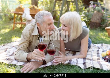 Liebend verheiratetes Seniorenpaar trinkt draußen Wein, liegt auf einer Decke, während es in ihrem Garten picknickt Stockfoto