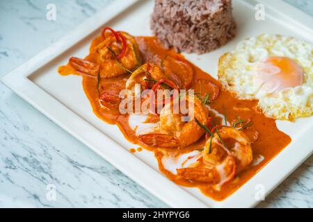 Chu Chee Kung oder getrocknetes rotes Kurry mit Garnelen, traditionelle thailändische Speisegarnelen getrocknetes rotes Kurry auf weißem Teller mit einem Spiegelei und Bio-Reis, auf dem gebraten wird Stockfoto