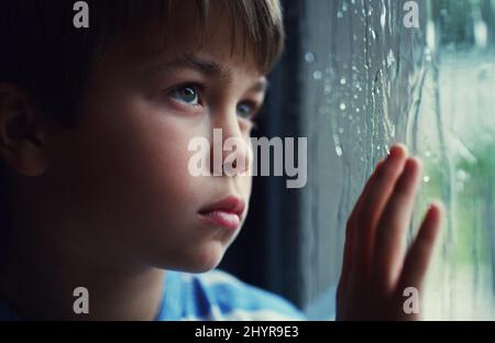 Wenn die Natur Ihren Plan zum Spaß sabotiert. Aufnahme eines traurigen Jungen, der den Regen durch ein Fenster zu Hause beobachtete. Stockfoto