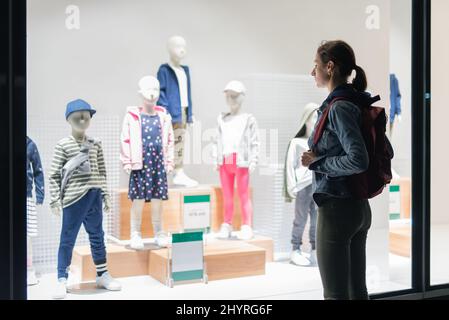 Frau steht vor dem Kindergeschäft und schaut sich die Kleidung auf den Schaufensterpuppen an Stockfoto