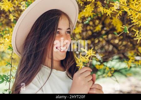 Glückliches kleines lächelndes Mädchen, das gelbe Blumen unter blühendem Forsythia-Busch im Frühlingsgarten hält. Kind trägt Hut Stockfoto