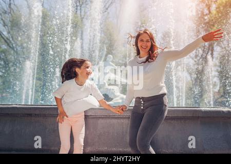 Muttertag. Junge Mutter und ihre Tochter haben Spaß am Brunnen im Sommerpark. Glückliche Familie springen und lachen Stockfoto