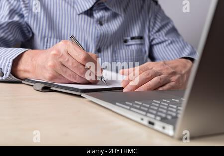 Mann, der mit einem Laptop am Tisch sitzt und mit einem Stift in einem Tagebuch schreibt. Nahaufnahme der Hände. Teilnahme an Online-Kursen, Prüfungsvorbereitung, Planungskonzept. Hochwertige Fotos Stockfoto